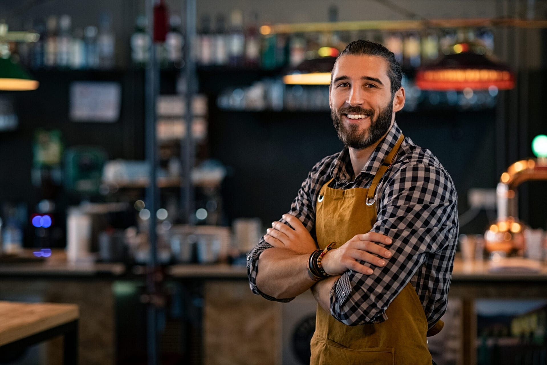 Happy,Satisfied,Bartender,With,Apron,Near,Counter,With,Crossed,Arms Bartender standing in front of the room with his arms crossed.
